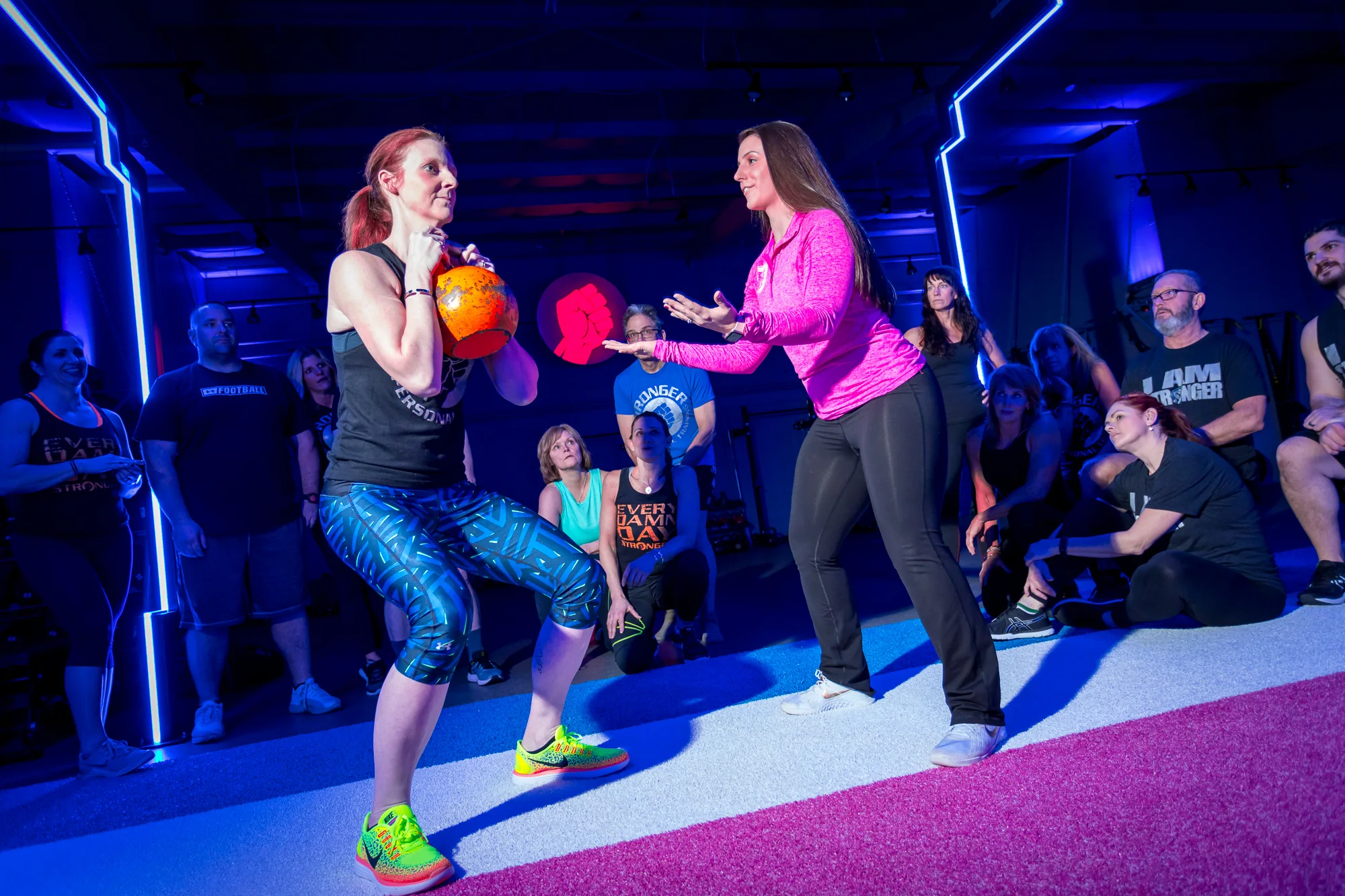 A woman practising a kettlebell lunge with a coach beside her and members nearby.
